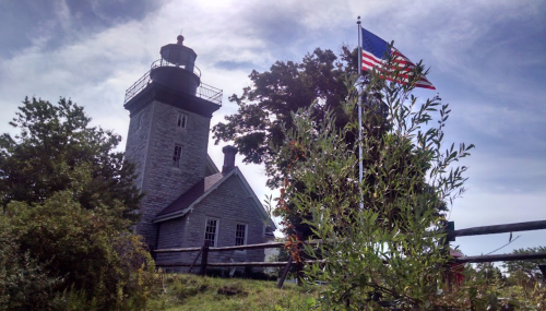 A historic lighthouse stands beside a waving American flag, surrounded by greenery under a blue sky.