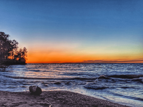 A serene beach at sunset, with vibrant orange and blue hues reflecting on the water and trees lining the shore.