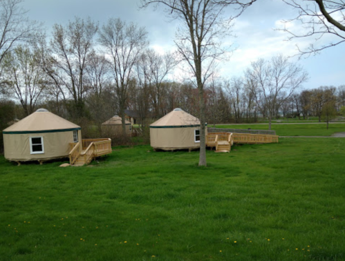 Two beige yurts with wooden decks sit on a grassy area, surrounded by trees under a cloudy sky.