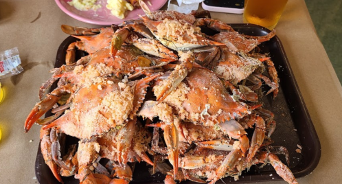 A large tray piled high with cooked crabs, covered in seasoning, with a side of food and a drink in the background.