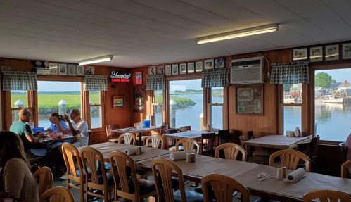 Cozy restaurant interior with wooden decor, tables set for dining, and a view of a river and boats outside.