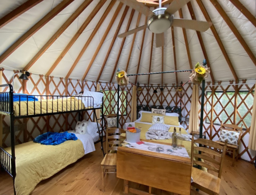 Cozy yurt interior with bunk beds, a dining table, and decorative pillows, featuring wooden beams and natural light.