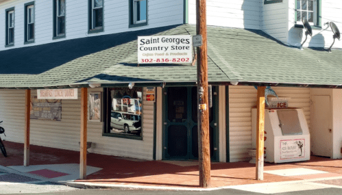 A small country store with a green roof and sign, featuring a phone number and ice for sale outside.