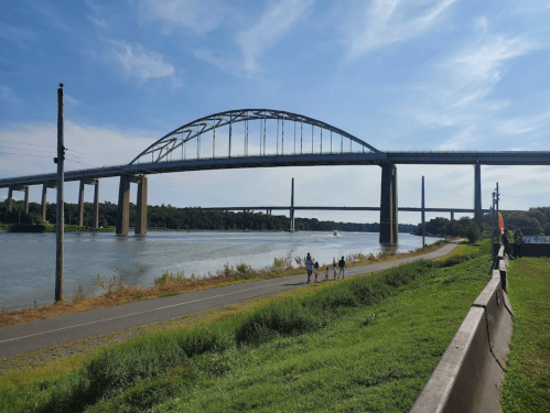 A wide river with a large arch bridge overhead, surrounded by greenery and a walking path along the water's edge.
