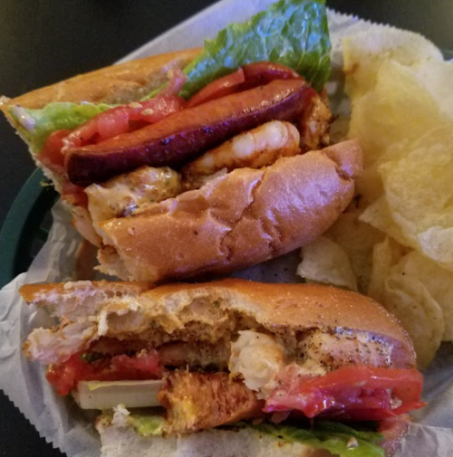 A close-up of a sandwich with lettuce, tomato, and sausage, served with a side of potato chips.