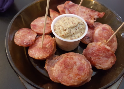 A plate of sliced sausage with toothpicks and a small cup of mustard in the center.