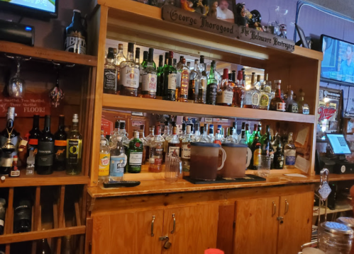 A wooden bar shelf displaying a variety of liquor bottles and glasses, with a TV in the background.