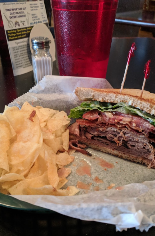 A hearty sandwich with layers of meat, lettuce, and tomato, served with a side of potato chips and a drink.