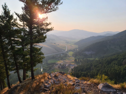 A scenic view of a valley at sunset, framed by trees, with rolling hills and a winding road below.