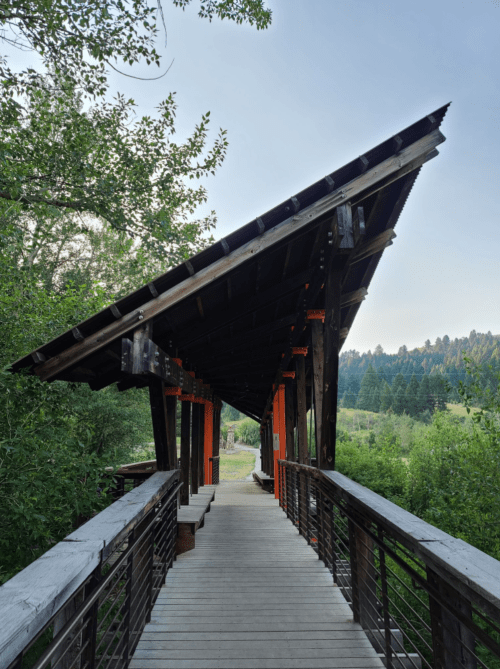A wooden bridge with a sloped roof leads into a lush green landscape, surrounded by trees and hills.