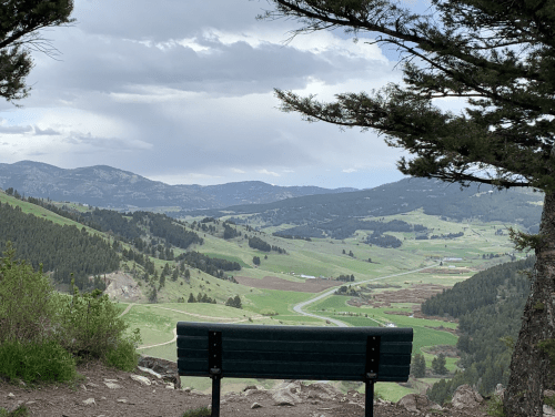 A green bench overlooks a scenic valley with rolling hills and distant mountains under a cloudy sky.