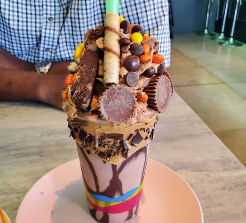 A decadent milkshake topped with chocolate, candy, and cookies, served in a colorful glass on a pink plate.