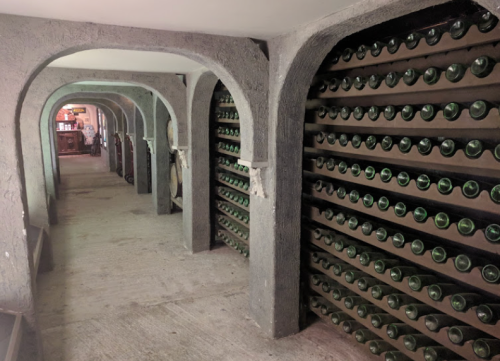 A dimly lit wine cellar with arched walls, featuring rows of green bottles on shelves.