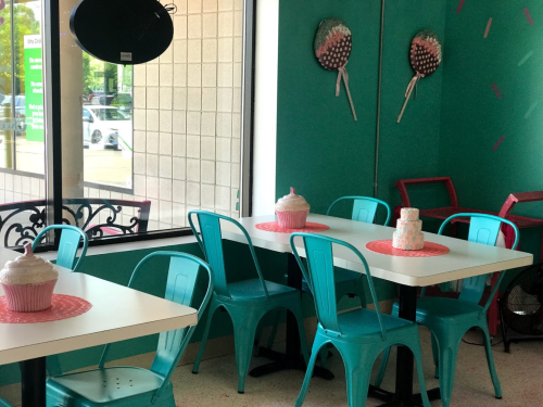 Bright café interior with teal chairs, white tables, and decorative cupcakes on each table against a green wall.