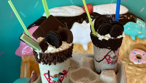 Two colorful milkshakes topped with whipped cream, chocolate, and cookies, served in a tray against a playful background.