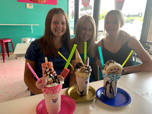 Three girls smile at a table with colorful milkshakes topped with sweets and bright straws in a vibrant ice cream shop.