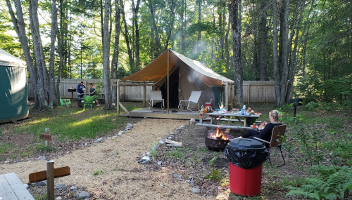 A campsite with a tent, picnic table, fire pit, and chairs surrounded by trees and a wooden fence.