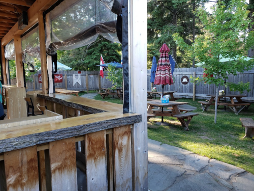 Outdoor seating area with picnic tables, umbrellas, and a bar under a wooden structure surrounded by trees.