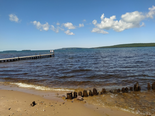 A serene beach scene with a wooden pier, calm water, and distant hills under a blue sky with fluffy clouds.