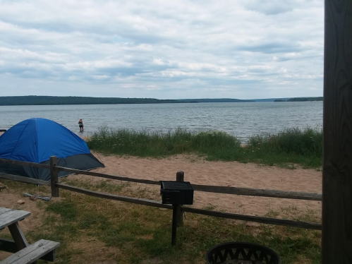 A tent by a sandy beach with a person standing in the water, surrounded by grass and a cloudy sky.