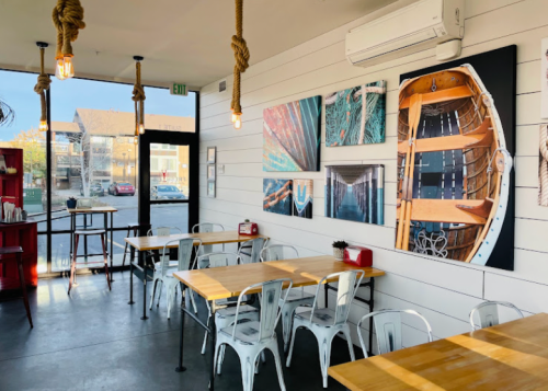 Bright café interior with wooden tables, metal chairs, and nautical-themed artwork on the walls. Large windows let in natural light.