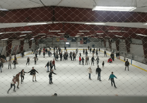 A busy indoor ice skating rink filled with skaters of all ages, viewed through a netted barrier.