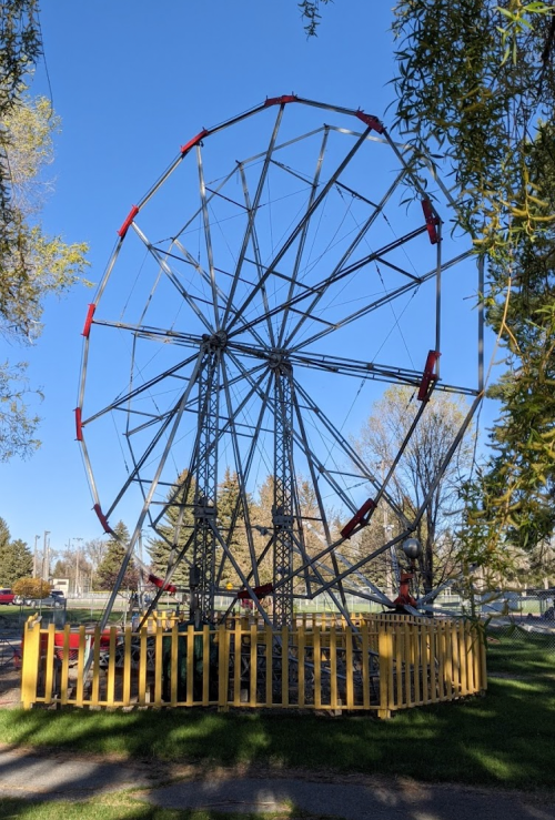 A large, metal Ferris wheel stands in a park, surrounded by a wooden fence and trees under a clear blue sky.