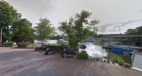A serene lakeside view with boats docked, surrounded by trees and greenery under a cloudy sky.