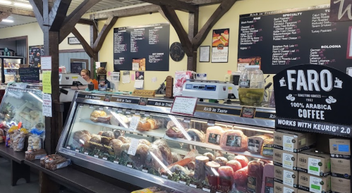 A bustling deli counter with various meats, cheeses, and baked goods, featuring a menu board in a rustic market setting.