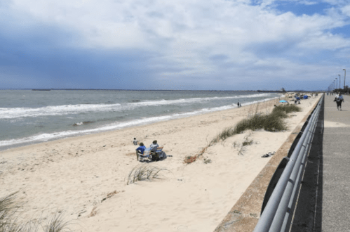 A sandy beach with people sitting and walking along the shore, under a partly cloudy sky. Waves gently lap at the sand.