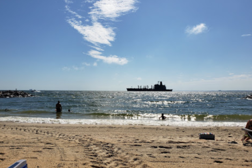 A beach scene with people swimming and a silhouette of a ship on the horizon under a clear blue sky.