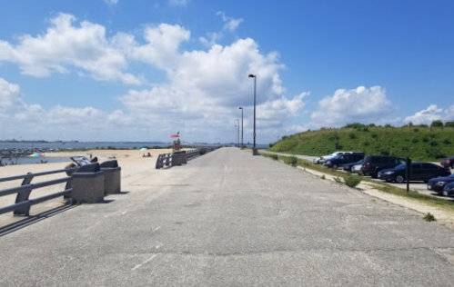 A wide, empty walkway along the beach with parked cars and a clear blue sky dotted with clouds.