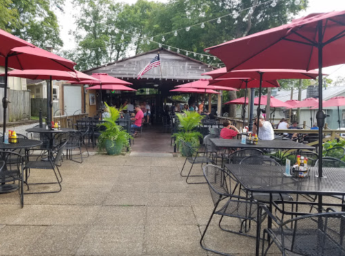 Outdoor dining area with black tables, red umbrellas, and greenery, featuring a rustic building and an American flag.