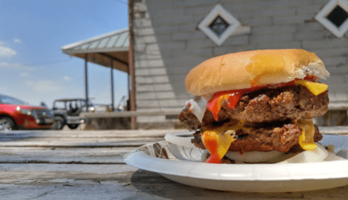 A close-up of a double cheeseburger with ketchup and mayonnaise on a paper plate, set on a wooden table outdoors.