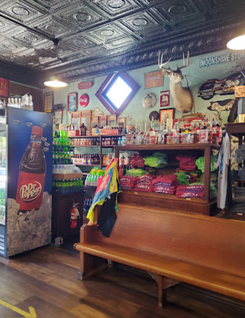 A rustic store interior with shelves of drinks, snacks, and colorful shirts, featuring a mounted deer head on the wall.