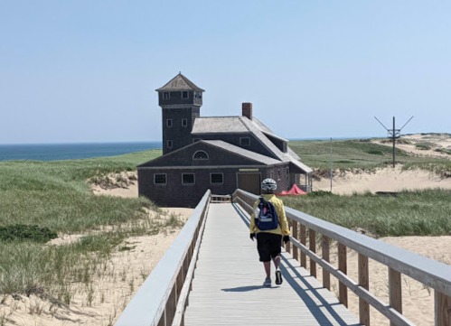 A person walks along a wooden boardwalk towards a beach house, with sand dunes and ocean in the background.