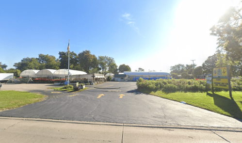 A sunny view of a garden center entrance with green grass, a flagpole, and large greenhouses in the background.