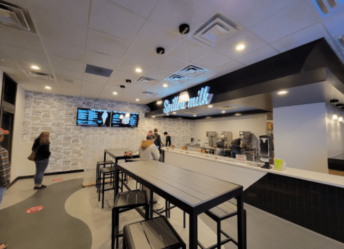 Interior of a modern café with a menu board, seating area, and customers ordering at the counter. Bright and inviting atmosphere.