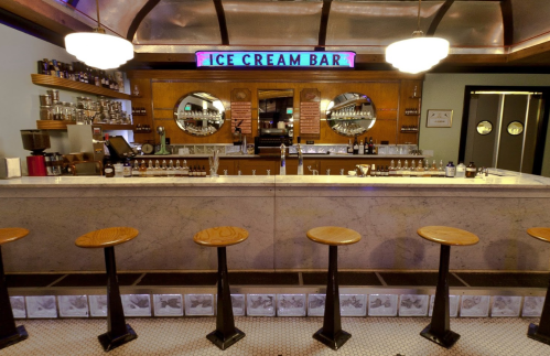 A vintage ice cream bar with a marble counter, wooden stools, and retro signage, featuring mirrors and a well-stocked shelf.
