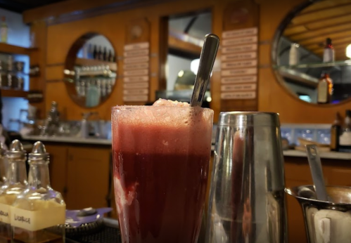 A frosty drink in a glass with a spoon, set against a bar with bottles and mirrors in the background.