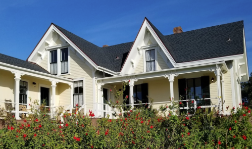 A charming two-story house with a gabled roof, surrounded by vibrant red flowers under a clear blue sky.