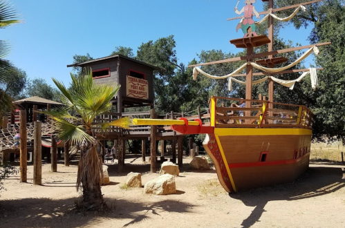 A colorful pirate ship playground surrounded by trees and rocks, with a wooden structure in the background.