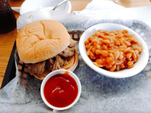A pulled pork sandwich on a bun with a side of baked beans and a small cup of barbecue sauce.