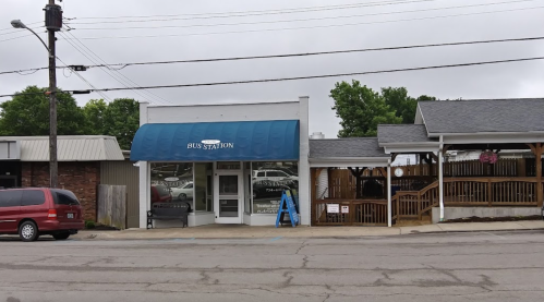 A small bus station with a blue awning, located on a street with nearby buildings and a wooden structure.