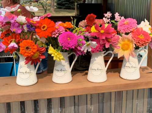 Four white pitchers filled with vibrant, colorful flowers sit on a wooden counter, creating a cheerful display.