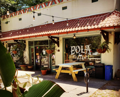 A charming café with a red-tiled roof, outdoor seating, and potted plants, set in a sunny, inviting atmosphere.