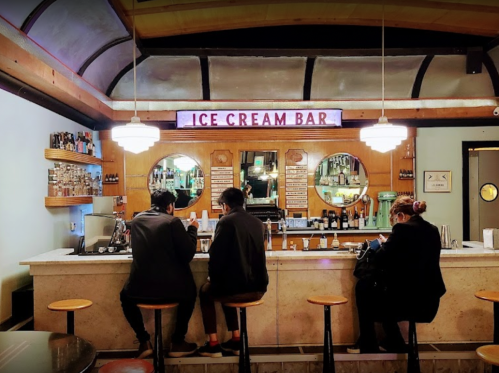 Two people sit at a vintage ice cream bar, with a wooden counter and retro decor, under warm lighting.