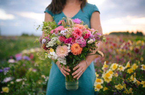 A person in a teal dress holds a large bouquet of colorful flowers in a vibrant field.