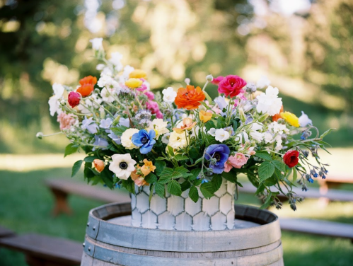 A vibrant floral arrangement in a patterned vase atop a wooden barrel, set in a lush outdoor environment.