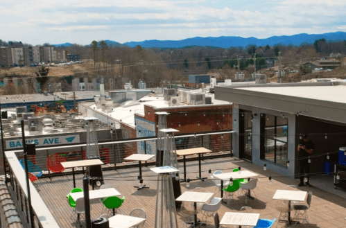 Rooftop view with tables and chairs, overlooking a town and mountains in the background under a clear sky.